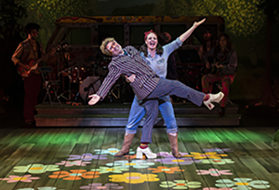 As You Like It, Shakespeare Theatre Company, Touchstone Audry in denem shirt and pants and brown boots hods Touchstone wearing striped jacket and platform shoes on the flower lit stage, with the band in the background shadows.